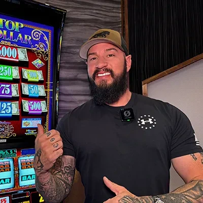 A bearded man in a baseball cap standing beside a slot machine, giving a thumbs-up gesture.