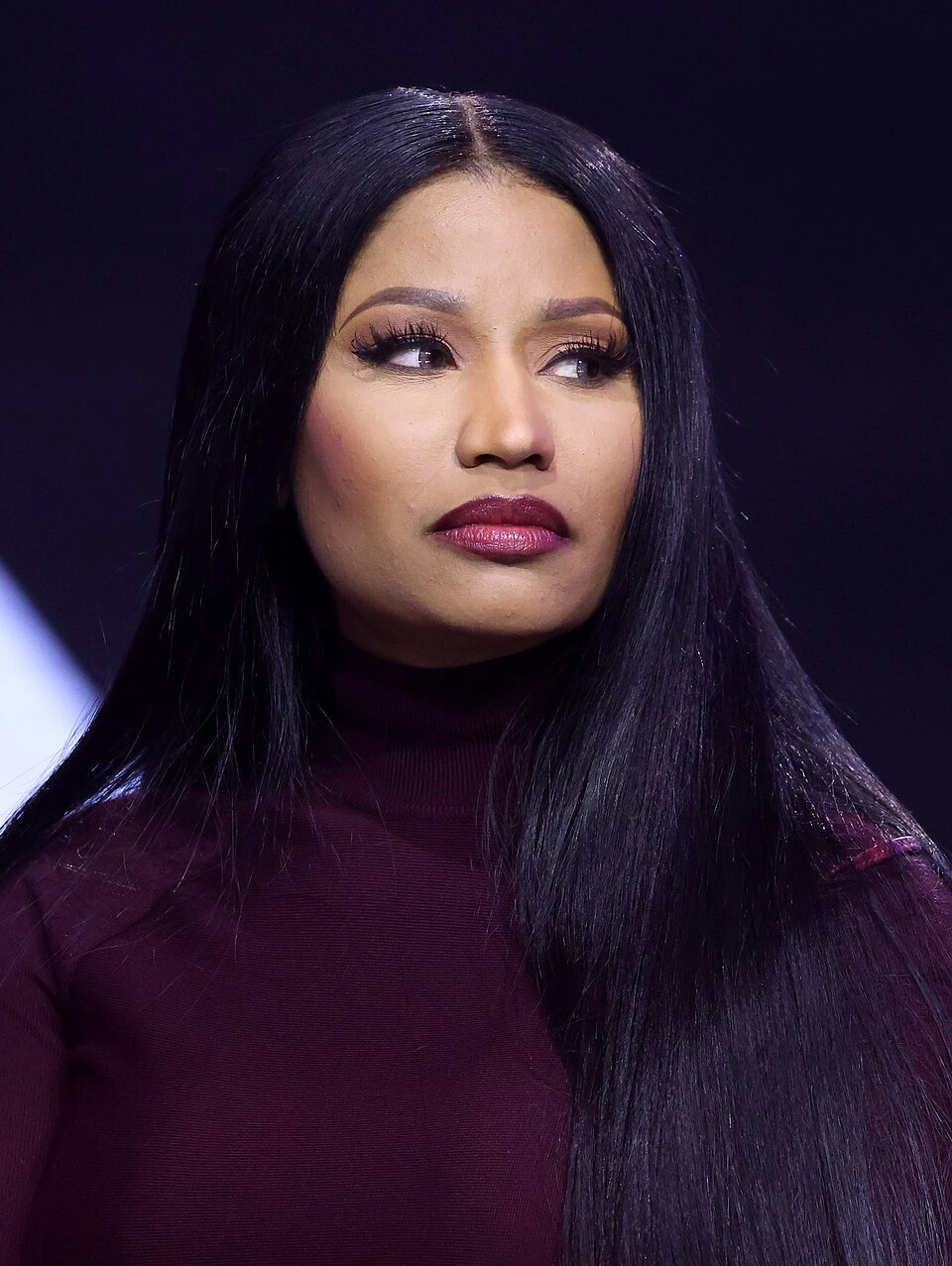 Nicki Minaj in a studio-style portrait, wearing a dark purple top with long straight black hair against a dark backgroun