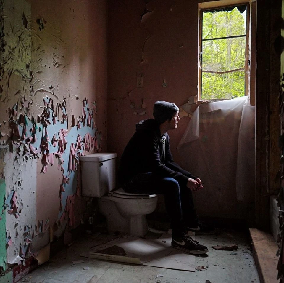 A person sitting on a toilet in a decaying room with peeling paint, looking out a window in an abandoned building.