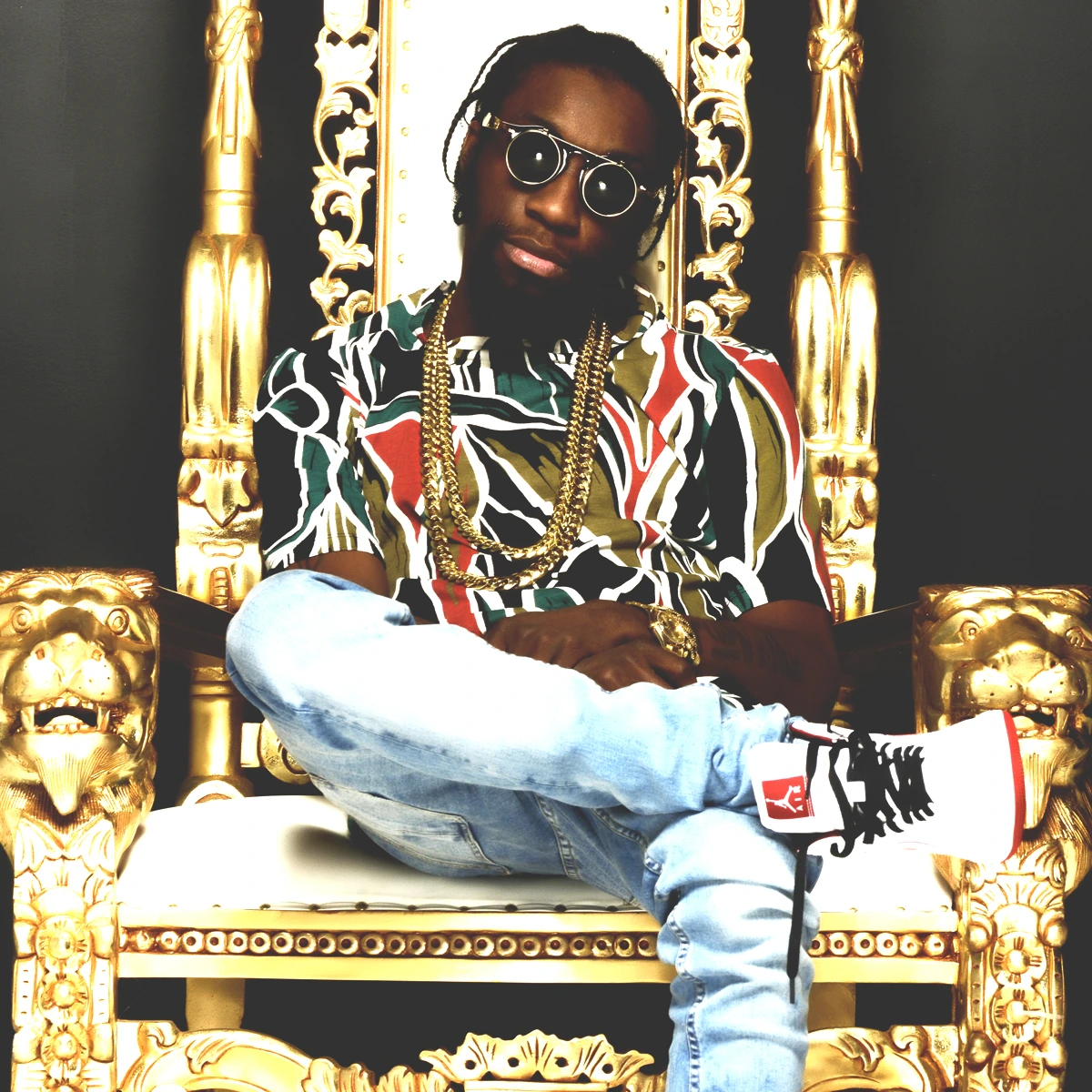 RajahWild seated on an ornate gold throne, wearing sunglasses, chains, and a patterned shirt.