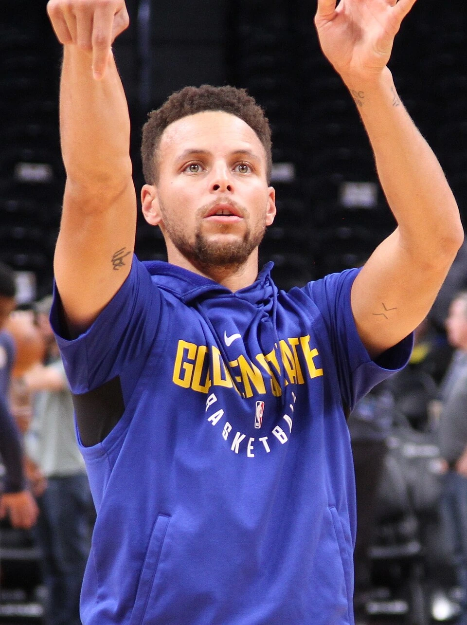 Stephen Curry warming up in a Golden State Warriors hoodie, arms raised on the court
