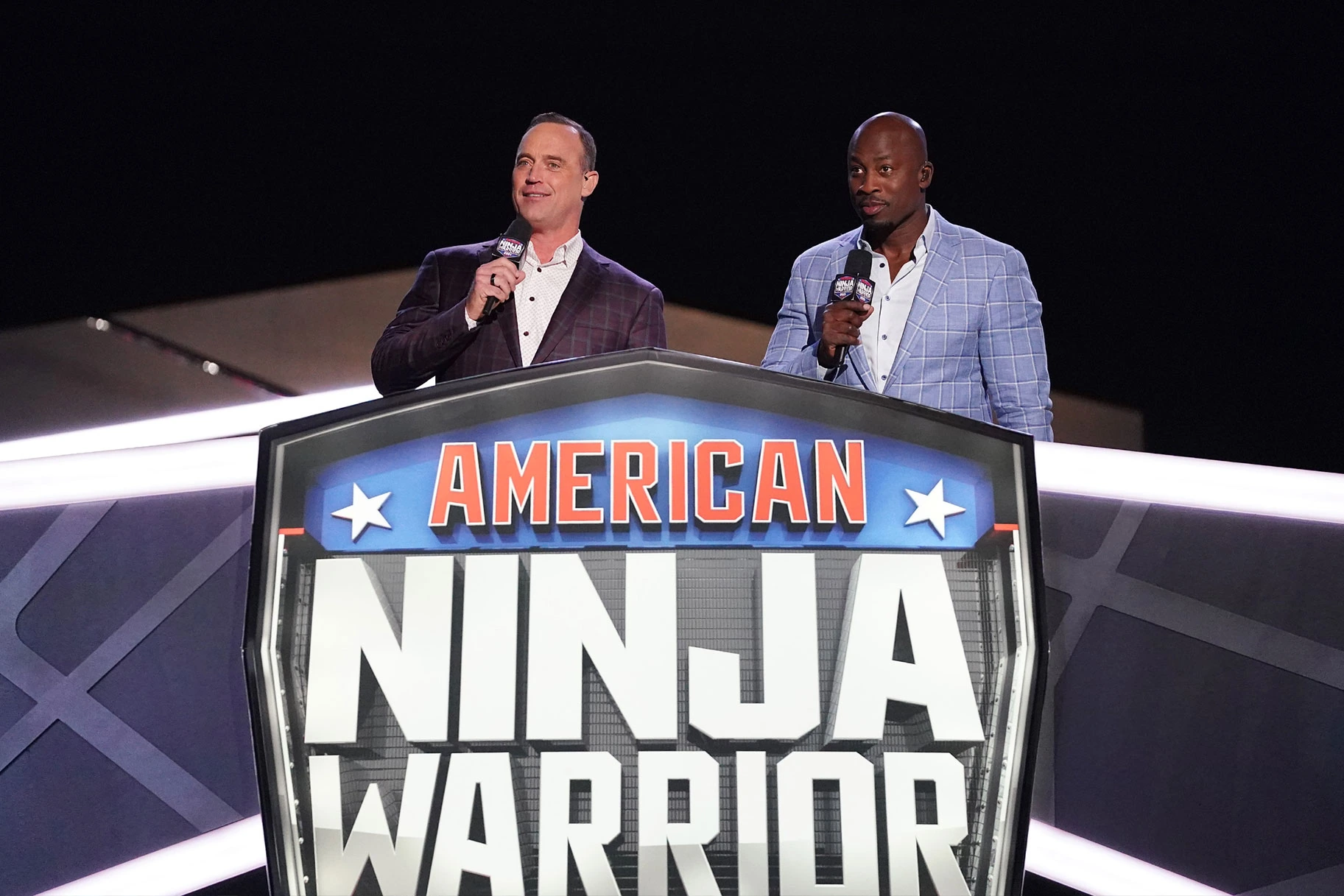 Matt Iseman standing on stage holding a microphone beside the American Ninja Warrior sign